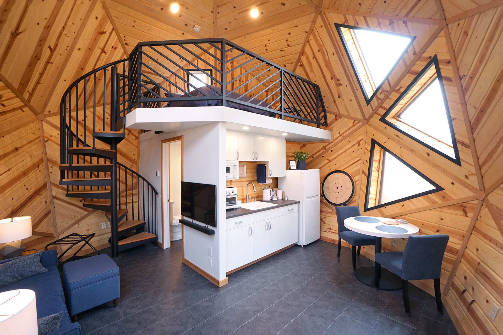 Wide view of the mini dome interior showcasing the sleeping loft, kitchenette, dining area, couch, bathroom, spiral staircase, slate flooring, and knotty pine walls.